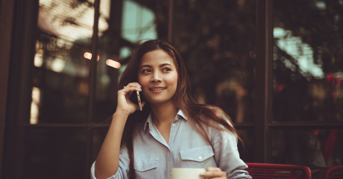 Young woman in casual shirt talking on phone and holding coffee at an outdoor café table.